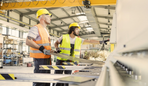 Two factory workers wearing hard hats and high-visibility vests handle a large sheet of metal beside industrial machinery in a brightly lit manufacturing facility. One worker points toward the machine while metal shavings fly from the cutting area.