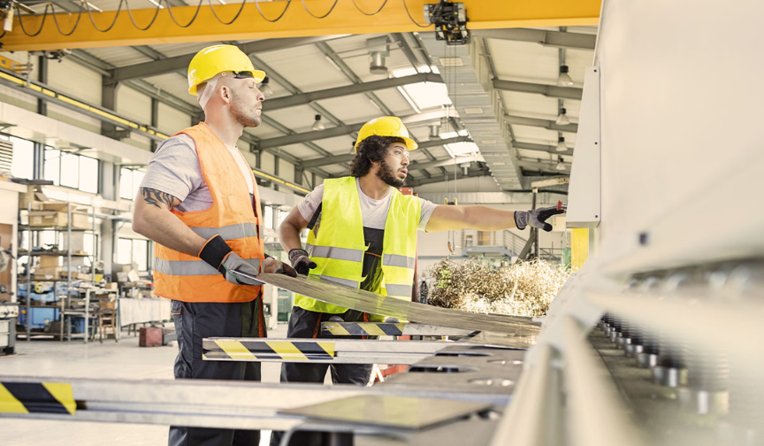 Two factory workers wearing hard hats and high-visibility vests handle a large sheet of metal beside industrial machinery in a brightly lit manufacturing facility. One worker points toward the machine while metal shavings fly from the cutting area.